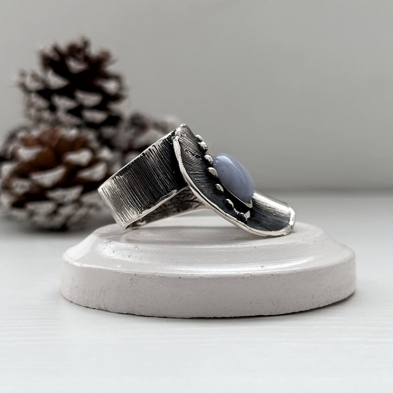 Puede incluir: Un anillo de plata con una banda texturizada y una piedra preciosa azul claro engastada en un engaste de plata. El anillo est&aacute; sobre un expositor blanco.