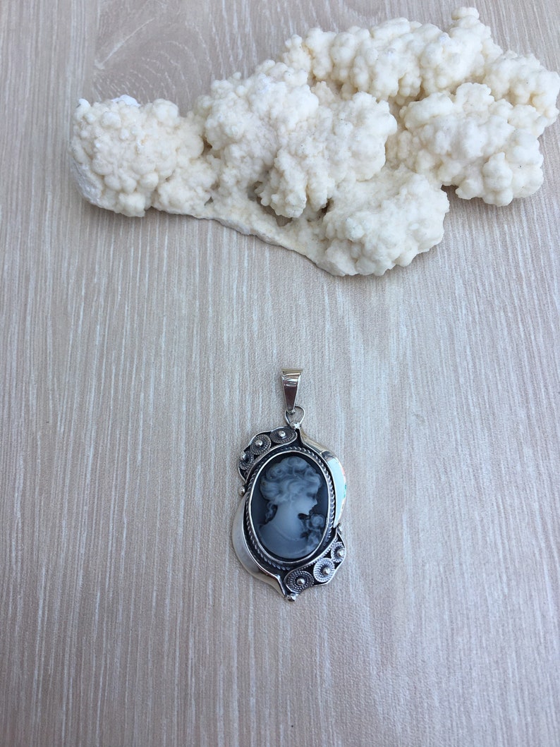 May include: A silver cameo pendant with a black and white portrait of a woman. The pendant has a detailed design with floral accents.