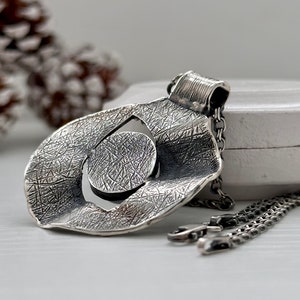 Puede incluir: Un collar de plata con un colgante de dise&ntilde;o circular y texturado. El colgante tiene un c&iacute;rculo m&aacute;s peque&ntilde;o cortado en el centro. El collar est&aacute; en una cadena de plata.