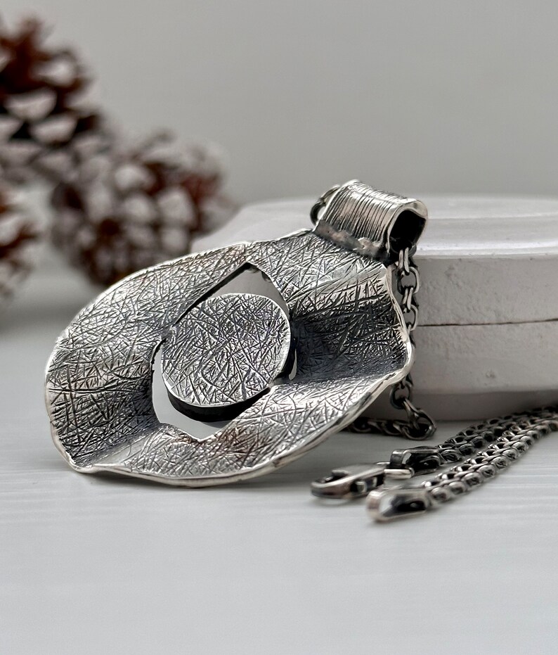 Puede incluir: Un collar de plata con un colgante de dise&ntilde;o circular y texturado. El colgante tiene un c&iacute;rculo m&aacute;s peque&ntilde;o cortado en el centro. El collar est&aacute; en una cadena de plata.