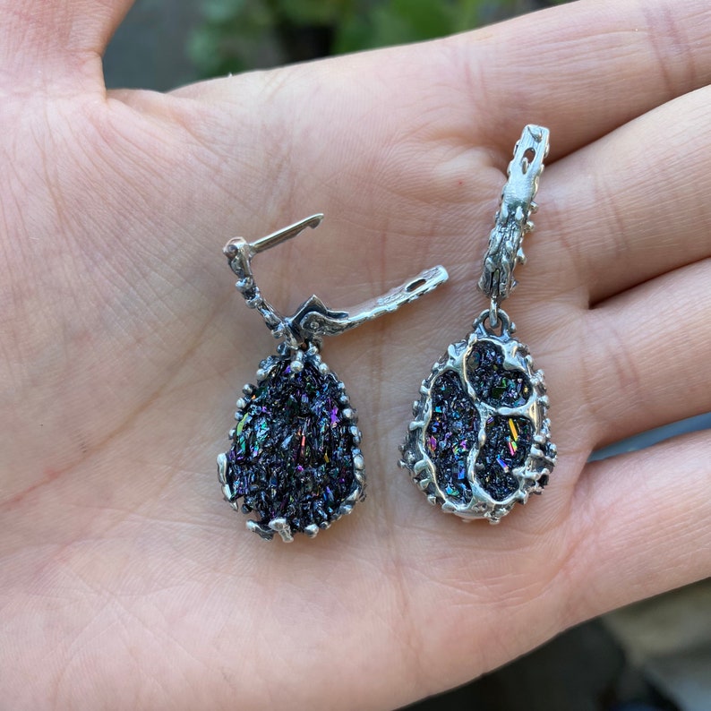 May include: A pair of silver earrings with iridescent black teardrop-shaped stones. The stones are set in a silver frame and have a rough, textured surface.