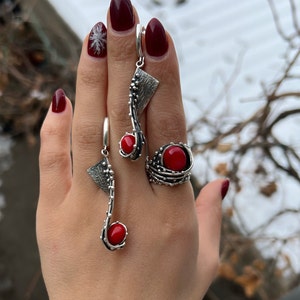 Puede incluir: Un conjunto de pendientes de plata y un anillo con piedras de coral rojo. Los pendientes son largos y colgantes, con un diseño geométrico. El anillo es una banda con una piedra de coral grande engastada en el centro.