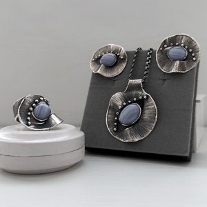 Puede incluir: Un conjunto de joyas de plata con un colgante de &aacute;gata azul, pendientes y anillo. Las joyas se muestran sobre una superficie de terciopelo gris.