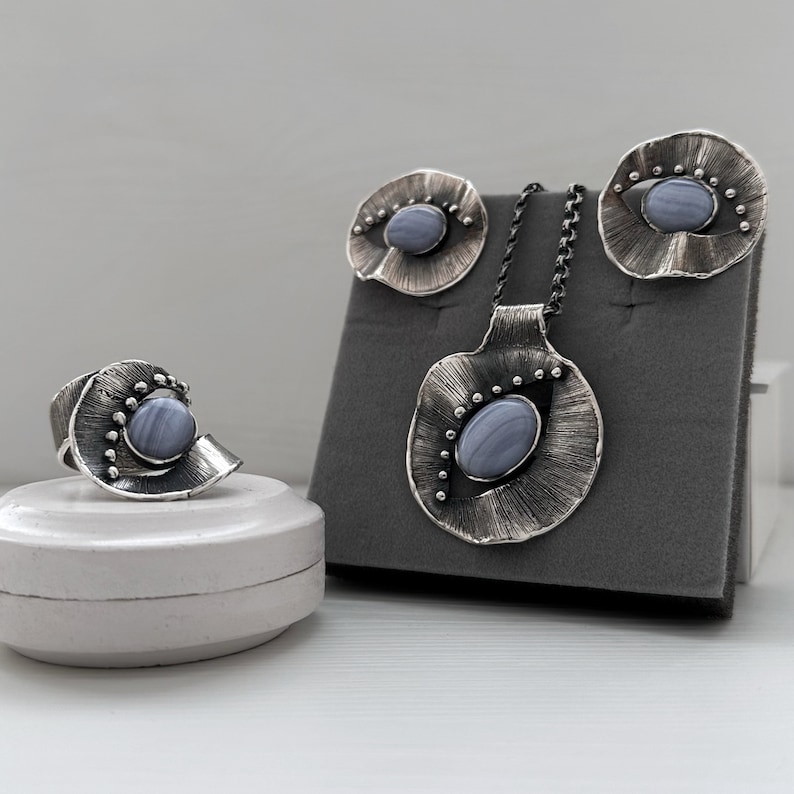 Puede incluir: Un conjunto de joyas de plata con un colgante de &aacute;gata azul, pendientes y anillo. Las joyas se muestran sobre una superficie de terciopelo gris.