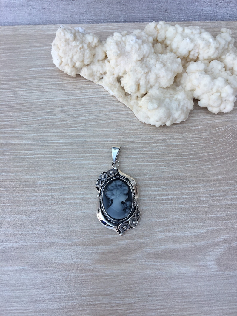 May include: A silver pendant with a black cameo featuring a woman's profile. The pendant has a detailed design with small silver beads and swirls.
