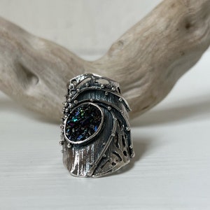 May include: A silver ring with a textured, organic design. The ring features a large, iridescent gemstone set in the center.