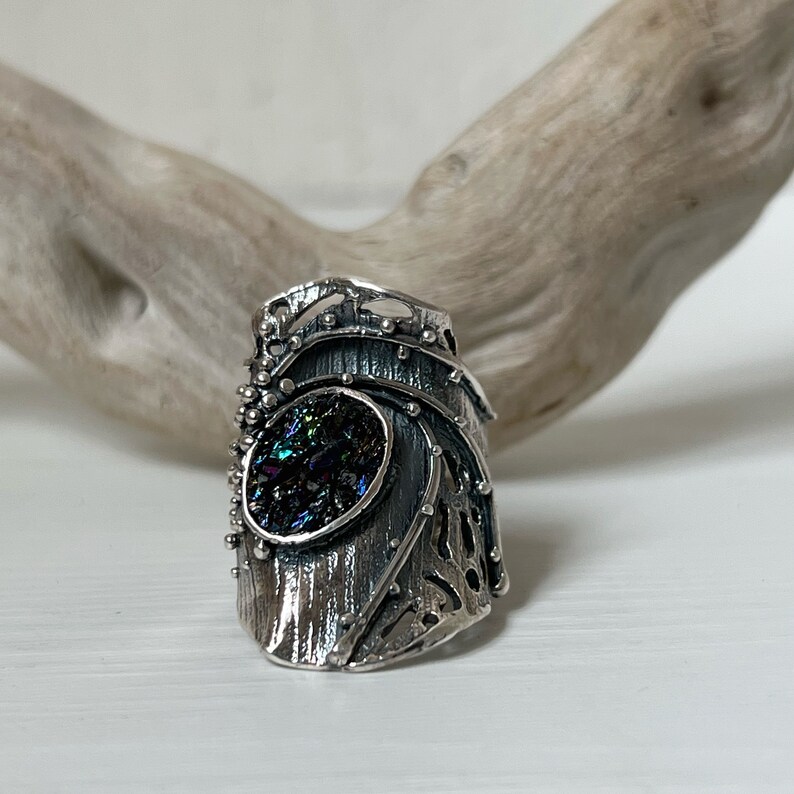 May include: A silver ring with a textured, organic design. The ring features a large, iridescent gemstone set in the center.