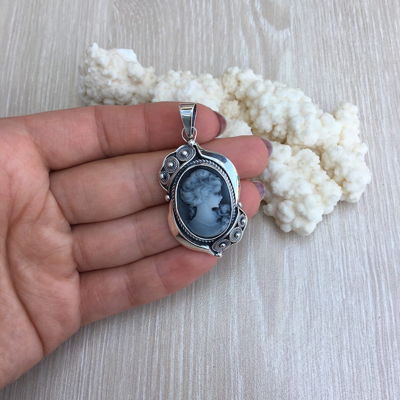 May include: A silver cameo pendant with a black and white portrait of a woman. The pendant has a detailed design with a rope border and small decorative circles.