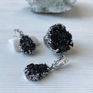 May include: A set of silver jewelry featuring iridescent black gemstone earrings, a ring, and a pendant. The jewelry is made with a rough, organic design.