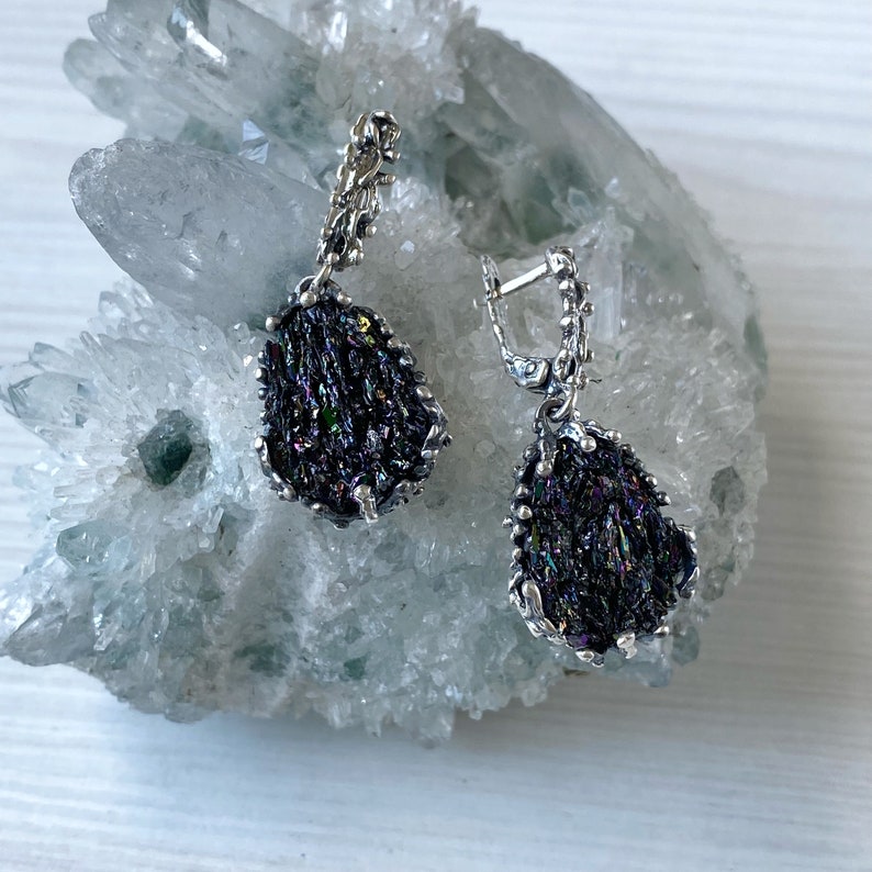 May include: A pair of silver earrings with iridescent black gemstone drops. The earrings are displayed on a white crystal cluster.