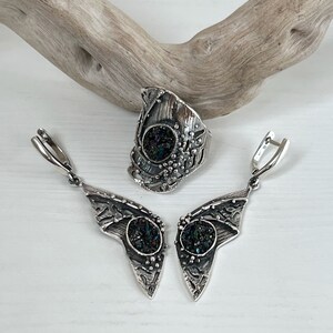 May include: A silver ring and earrings set with a black iridescent stone. The ring is a wide band with a textured design and the earrings are long and dangle.