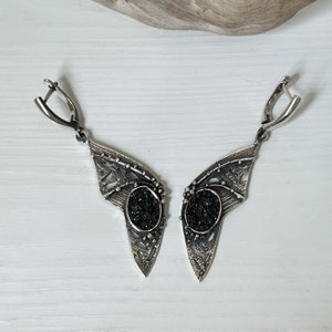 May include: A pair of silver earrings with a black, iridescent stone set in a delicate, intricate design. The earrings have a wing-like shape and dangle from a silver hook.