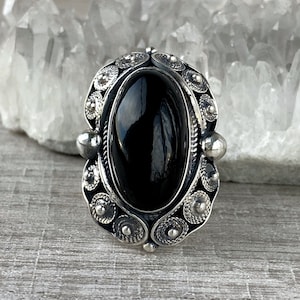 May include: A silver ring with an oval black onyx gemstone set in a filigree design.