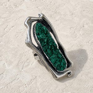 Puede incluir: Anillo plateado con un diseño orgánico y texturizado. El anillo presenta una piedra rectangular de color verde oscuro engastada en un marco. La piedra tiene una superficie rugosa e irregular, creando un contraste llamativo con el metal liso.
