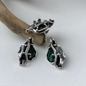 May include: A silver ring and earrings set featuring a green stone and a lizard design. The ring is open and the earrings are dangle style.