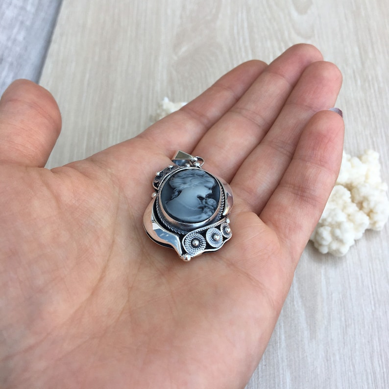 May include: A silver pendant with a black cameo featuring a woman's profile. The pendant has a detailed design with a swirl pattern and three round accents.