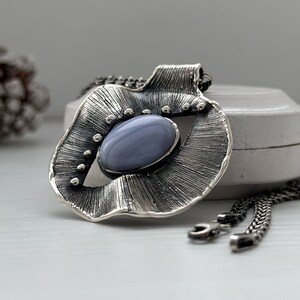 Puede incluir: Un collar de plata con un colgante que presenta una piedra de &aacute;gata azul engastada en un dise&ntilde;o texturizado en forma de abanico.