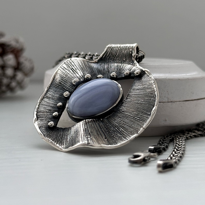 Puede incluir: Un collar de plata con un colgante que presenta una piedra de &aacute;gata azul engastada en un dise&ntilde;o texturizado en forma de abanico.