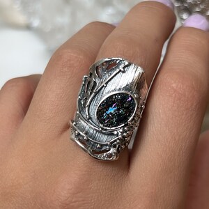 May include: A silver ring with an iridescent gemstone set in a detailed, textured design. The ring is wide and has a unique, organic shape.