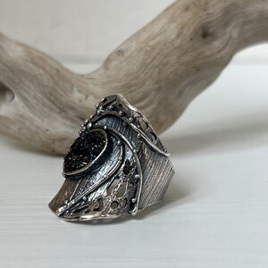 May include: A silver ring with a black stone set in a textured, geometric design. The ring is wide and has a unique, asymmetrical shape.