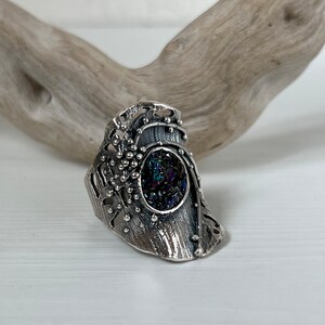May include: A silver ring with a detailed, textured design featuring a round, iridescent gemstone set in the center.