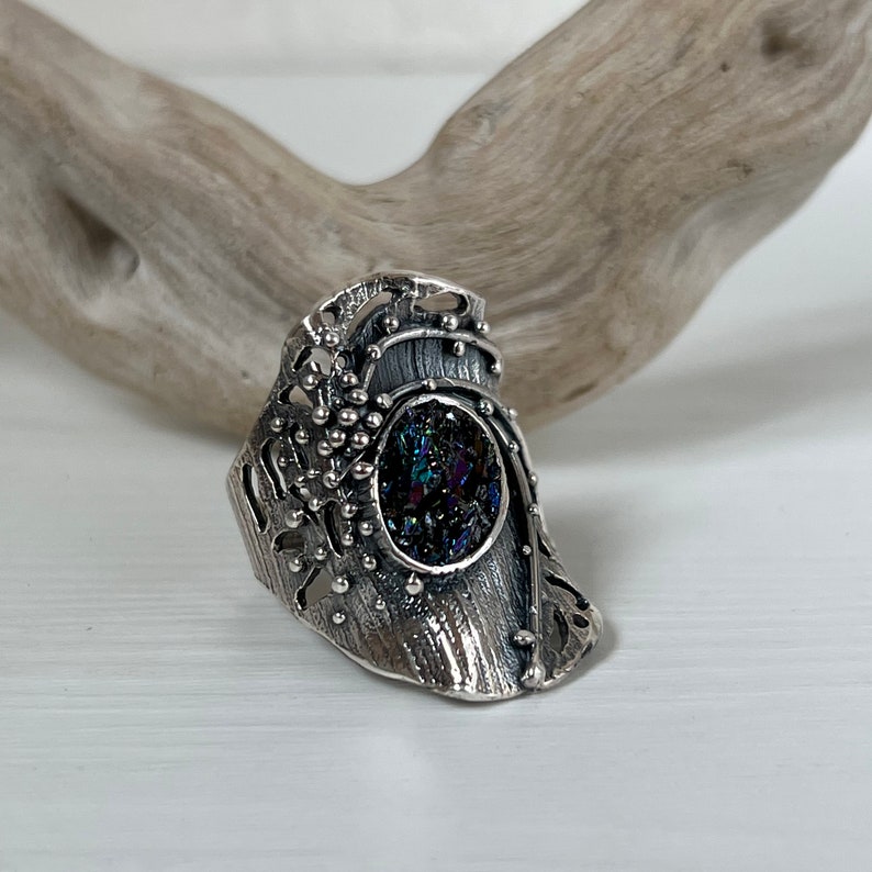 May include: A silver ring with a detailed, textured design featuring a round, iridescent gemstone set in the center.