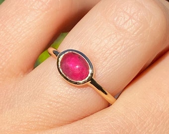 May include: A gold ring featuring an oval, vibrant red gemstone. The ring's band is a thin, polished gold, and the gemstone is set in a bezel setting. The ring is shown on a finger, with a blurred green and brown background.