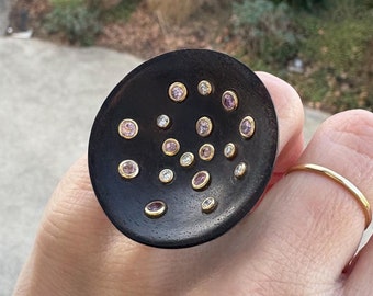 May include: A large, circular, dark brown ring with a concave surface featuring multiple small, round, bezel-set gemstones in shades of pink and clear white. The ring is worn on a finger alongside a thin, plain gold band on an adjacent finger.