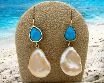 May include: A pair of gold earrings with turquoise and pearl drops. The earrings are displayed on a brown stand against a beach background.