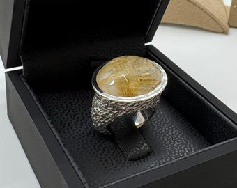 May include: A silver ring with a large, oval-shaped, clear gemstone with gold inclusions. The ring is in a black velvet box.