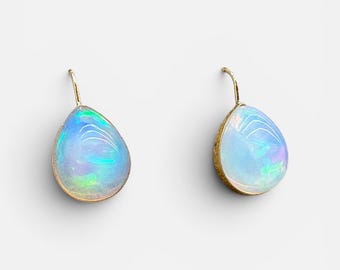 May include: A pair of teardrop-shaped earrings featuring iridescent opals set in gold-toned metal. Each earring has a curved hook wire for attachment, showcasing a smooth, polished surface with flashes of blue, green, and violet light reflections.