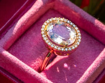 May include: A rose gold ring with a large, oval-cut gemstone. The gemstone is surrounded by a halo of small, clear stones. The ring is set in a pink velvet box, creating a luxurious presentation.