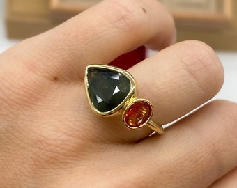 May include: A gold ring featuring a large, pear-shaped dark green gemstone and a smaller, oval-shaped orange gemstone. The stones are set in a smooth gold bezel setting on a thin gold band, creating a distinct two-stone design on the finger.