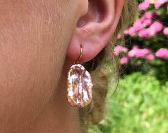 May include: A single drop earring featuring an irregular, elongated baroque pearl with a soft pink, iridescent luster. The pearl is suspended from a thin, polished gold-tone metal hook that passes through the earlobe, creating a dangling effect.