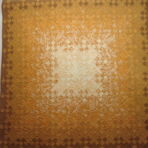 May include: A handmade quilt with a warm color palette. The center is a soft cream color, transitioning to shades of orange and brown. The border features a repeating diamond pattern in a darker brown.