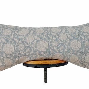 Könnte beinhalten: Ein rechteckiges Zierkissen mit einem floralen Muster in Hellblau und Off-White. Das Kissen liegt auf einem kleinen, runden Holzschemel mit schwarzem Metallsockel. Das Design des Kissens zeigt große, stilisierte Blumen.