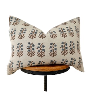 Op de afbeelding: Een beige linnen kussen met een bloemenpatroon in tinten bruin en blauw. Het kussen ligt op een houten kruk.