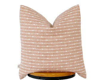 May include: A blush pink throw pillow with a white dotted stripe pattern. The pillow is sitting on a wooden stool.