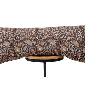 May include: A long, rectangular decorative pillow with a floral pattern in brown, beige, and teal. The pillow is resting on a small, round wooden surface supported by a black metal base.