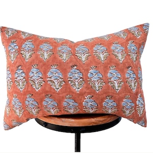 Puede incluir: Un cojín rectangular con un fondo naranja terracota y un patrón repetitivo de motivos florales en azul claro y beige dispuestos en filas horizontales a lo largo de toda la superficie de la funda de tela.