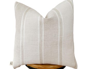 May include: A beige and white striped throw pillow with a subtle herringbone pattern. The pillow is sitting on a wooden stool.