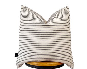 May include: A square pillow with a cream-colored background and horizontal black stripes. The pillow is placed on a wooden stool. A small black tag is visible on the lower left corner of the pillow.