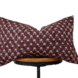 Puede incluir: Una almohada color burdeos con un estampado floral blanco. La almohada está sentada en un taburete de madera.