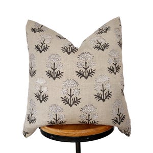 May include: A decorative throw pillow with a beige background and a repeating pattern of dark brown and white floral designs. The pillow is square and appears to be made of linen or a similar fabric. It is resting on a wooden stool.