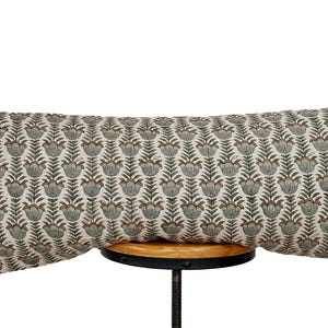 May include: A long, rectangular pillow with a white background and a repeating pattern of green and brown floral designs. The pillow is resting on a wooden stool.