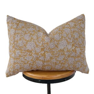 May include: A rectangular cushion with a floral pattern. The cushion has a mustard yellow background with white and grey floral designs. The cushion is resting on a wooden stool.