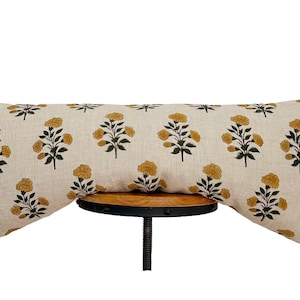 Könnte beinhalten: Ein langes, rechteckiges Kissen mit cremefarbenem Hintergrund und einem sich wiederholenden Muster aus gelben Blumen und dunkelgrünen Blättern. Das Kissen liegt auf einem kleinen, runden Holzschemel mit schwarzem Metallsockel.