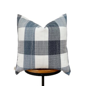 May include: A white and blue plaid throw pillow with a subtle textured fabric. The pillow is sitting on a wooden stool.