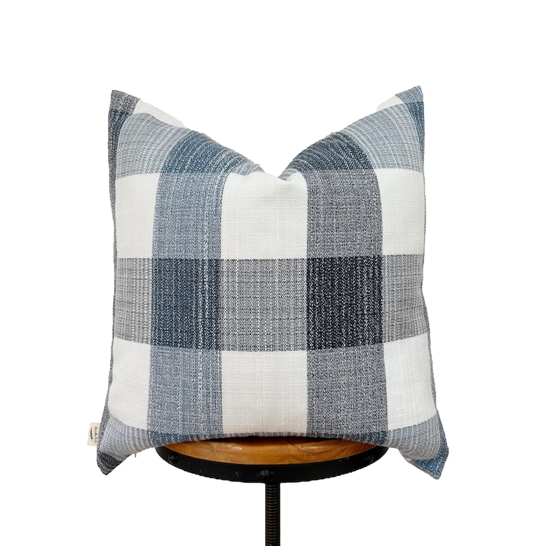 May include: A white and blue plaid throw pillow with a subtle textured fabric. The pillow is sitting on a wooden stool.