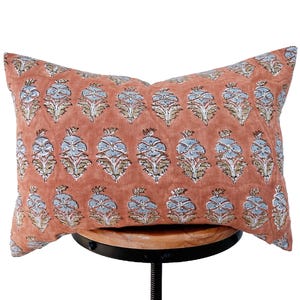 May include: A rectangular decorative pillow with a floral pattern. The pillow has a terracotta-colored background with a repeating pattern of blue and white flowers. The pillow is placed on a wooden stool.